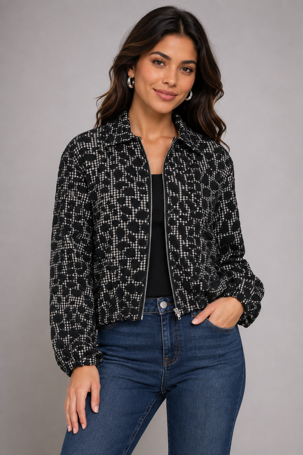A woman modeling a black and white patterned zip-up jacket over a black top and blue jeans, standing against a solid gray studio background.